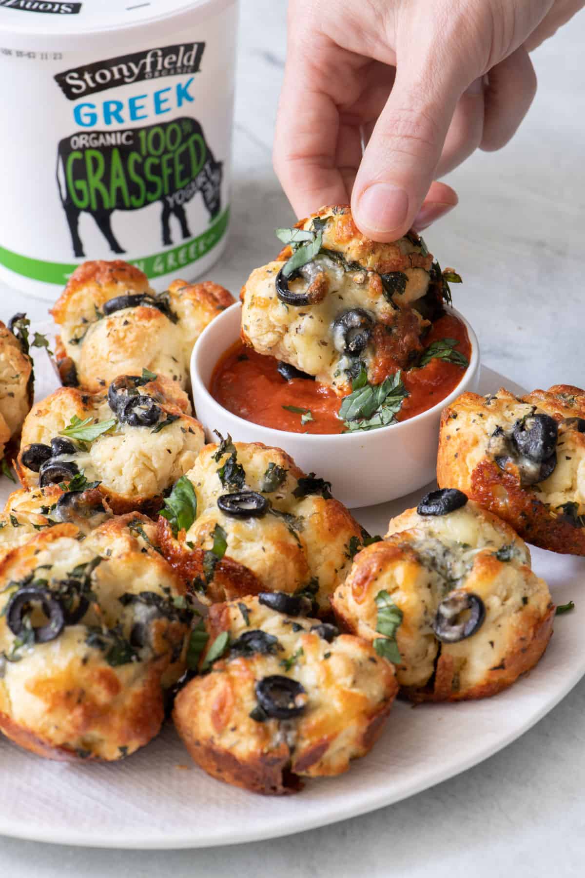 Hand dipping one pizza bite into pizza sauce with a tub of Stoneyfield Greek organic yogurt in background.