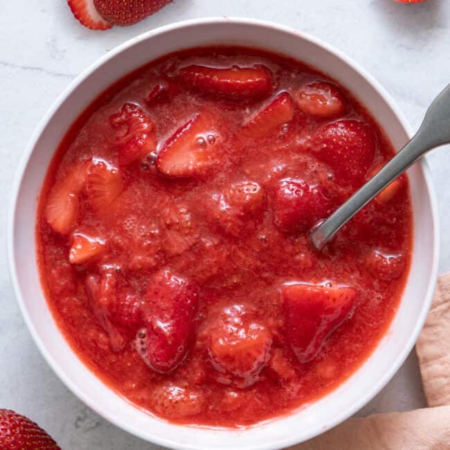 Homemade strawberry sauce in white bowl with a spoon and cut fresh strawberries around bowl.