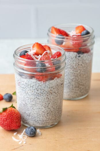 2 jars of chia seed pudding with chopped strawberries, blueberries, and just a few shreds of coconut on top with more fruit around jars.