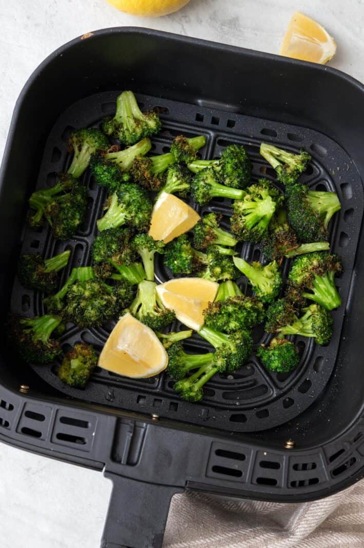 Air fried broccoli in an air fryer basket after cooking with lemon wedges.