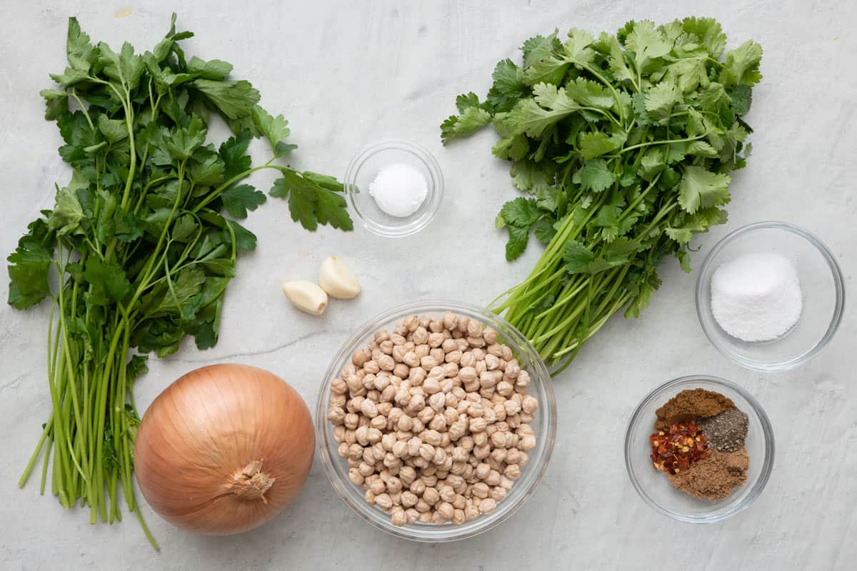 Ingredients for recipe before prepping: fresh cilantro and parsley, yellow onion, garlic cloves, salt, baking powder, spices, and dried chickpeas.