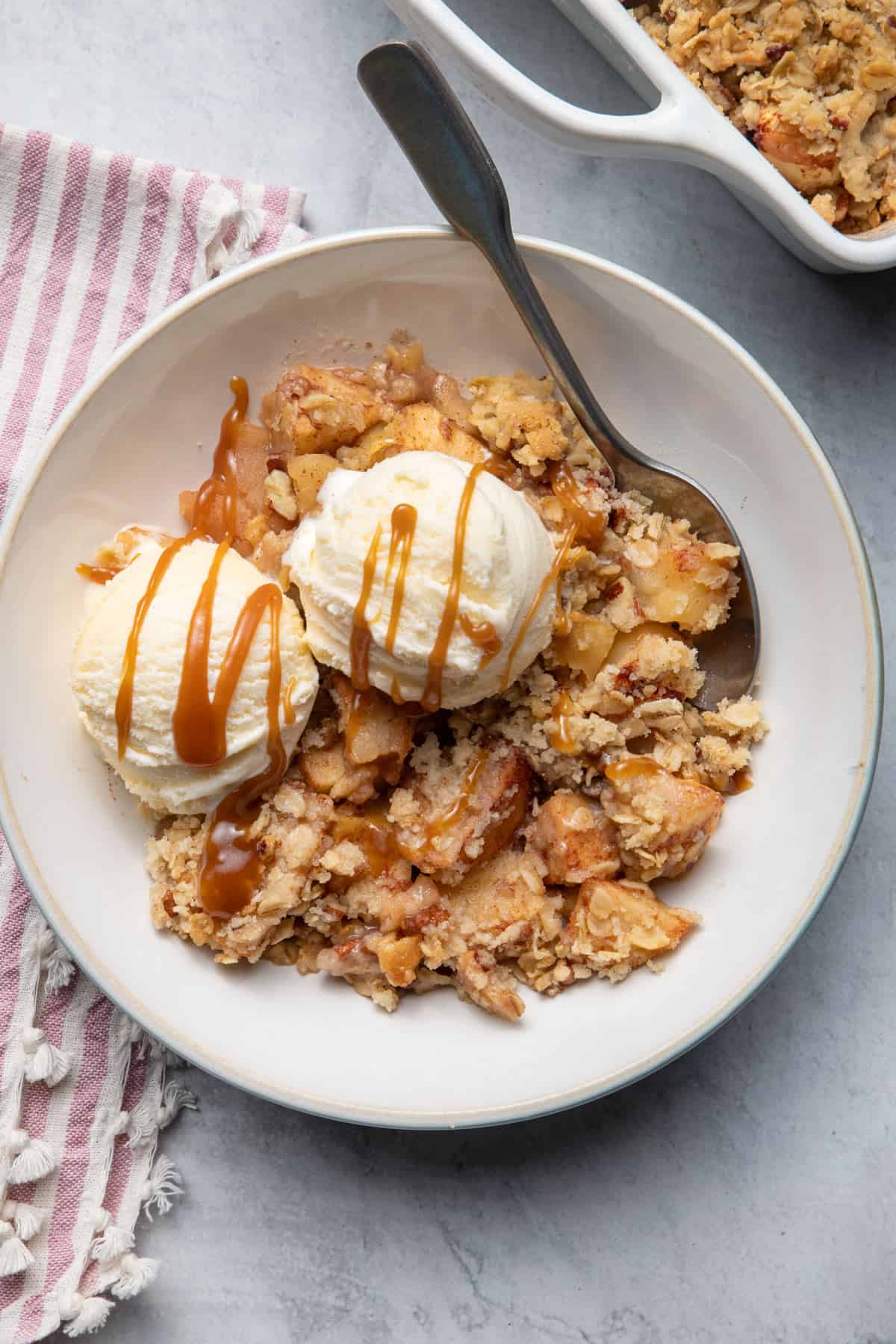 Apple crisp served with two scoops of vanilla ice cream and caramel sauce and spoon inside bowl