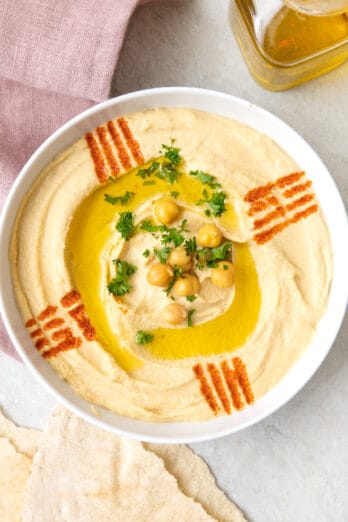 Bowl of authentic Lebanese hummus topped with olive oil, a few chickpeas, fresh chopped parsley, and a decorative lines made with paprike. Oil and pita nearby.