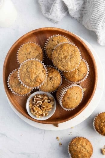 8 banana bread muffins stacked on a plate with a small dish of walnuts and a few extra muffins off plate.