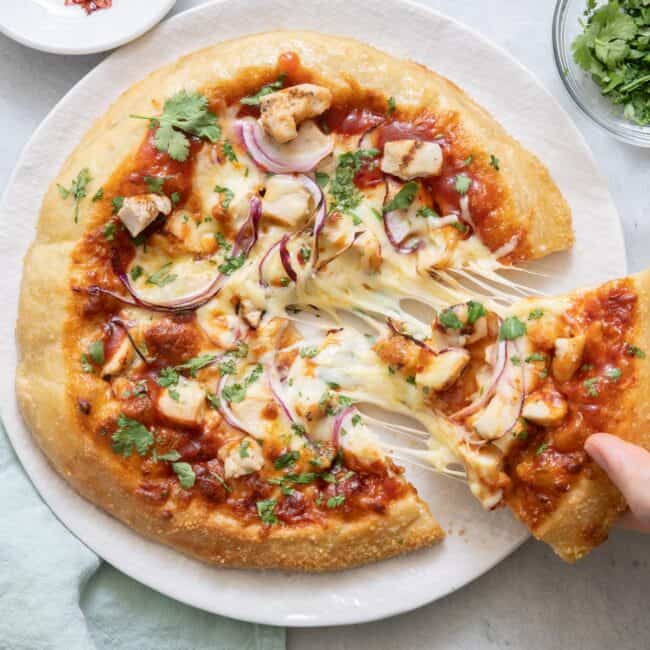 BBQ chicken pizza with a slice cut out being picked up from plate and showing melty cheese pulled from pizza.