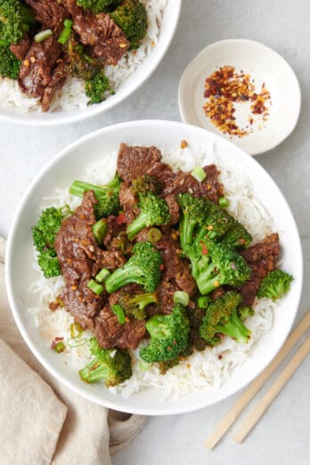 Beef and broccoli stir fry served in two bowls with a small dish of red pepper flakes and chop sticks nearby.