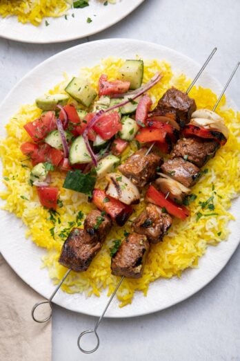 overhead shot of grilled beef kabobs on metal skewers on top of yellow rice and a side of vegetables