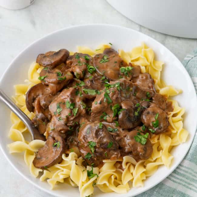 Plate of egg noodles topped with a serving of beef stroganoff, garnished with fresh parsley and a fork dipped inside bowl.