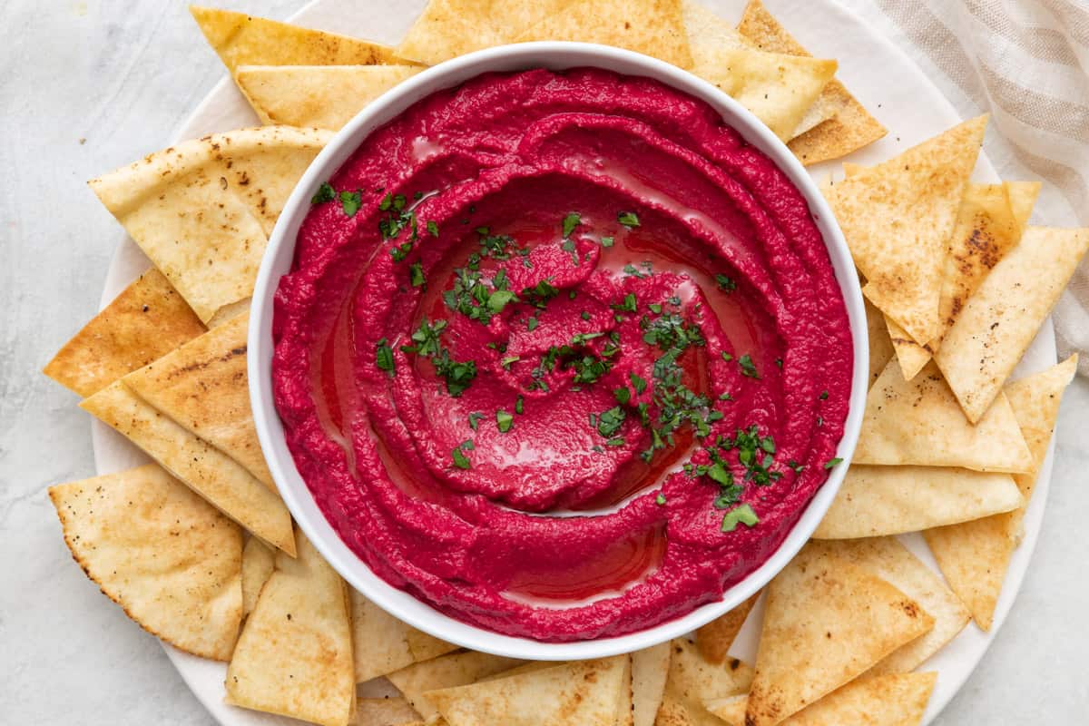 Beet hummus in a bowl garnished with fresh parsley that is set on a plate and surrounded with pita chips.