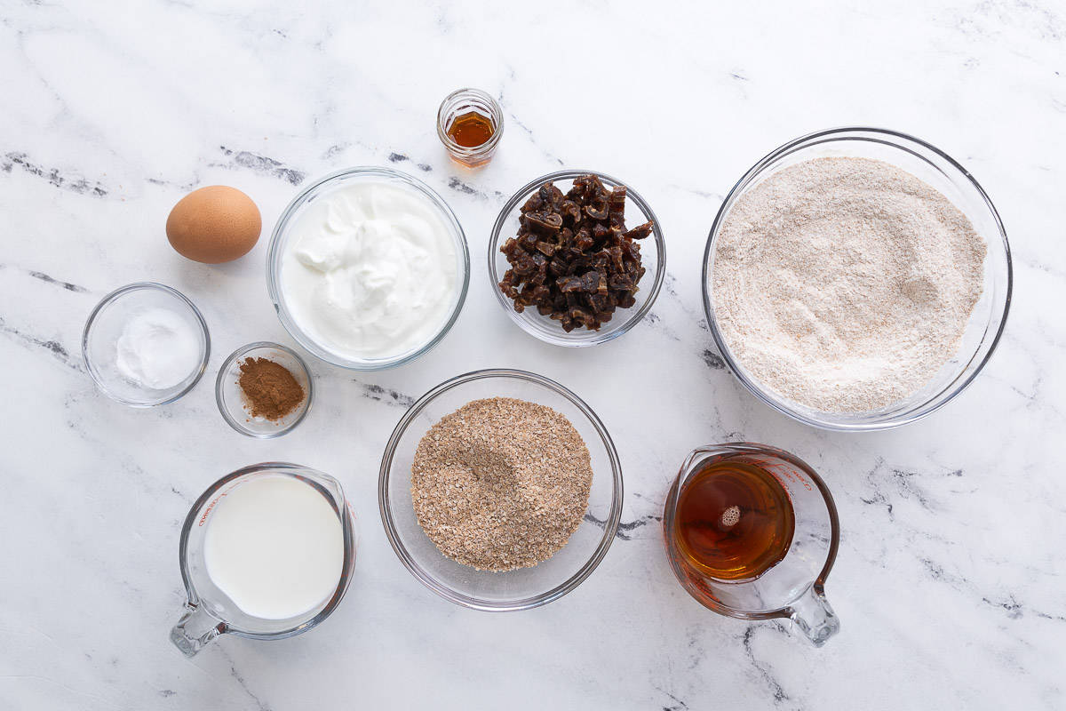Ingredients for recipe in individual bowls: baking powder and soda, cinnamon, egg, greek yogurt, milk, vanilla, chopped dates, wheat bran, maple syrup, and whole wheat flour.