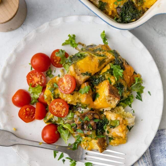 Plate of cheesy breakfast strata with halved cherry tomatoes garnished with fresh herbs and a fork with baking dish of recipe nearby.