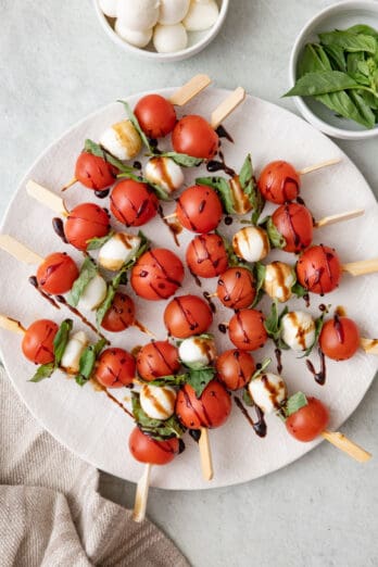 Caprese skewers on a large round plate with balsamic glazed drizzled over and a small dish of additional basil leaves and mozzarella balls nearby.