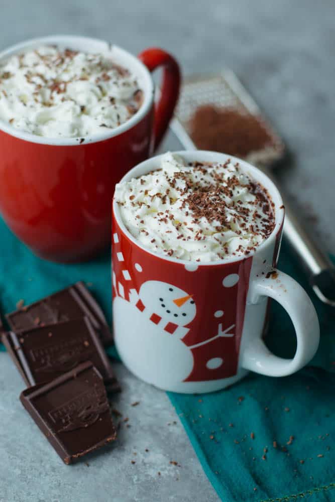 Vegan hot chocolate in two red mugs topped with whipped cream and shaved chocolate