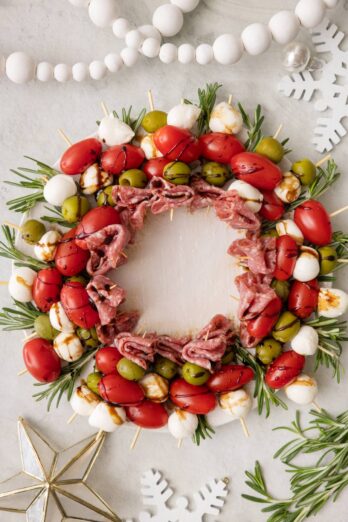 Charcuterie wreath full of individual skewers of tomatoes, salami, green olive, and mozzerella drizzled with balsamic glaze, garnished with rosemary sprigs and surrounded by white christmas decor.