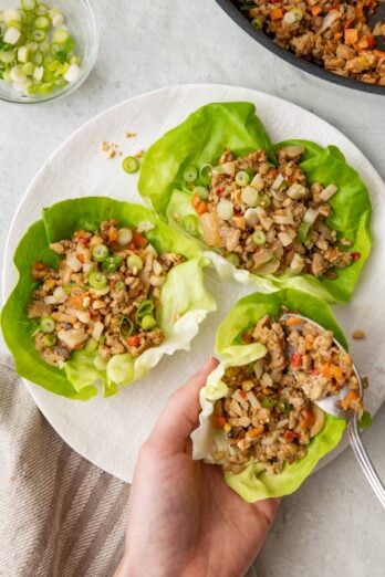 Chicken lettuce cups on a plate with a hand holding one and spooning in the filling to the lettuce cup.