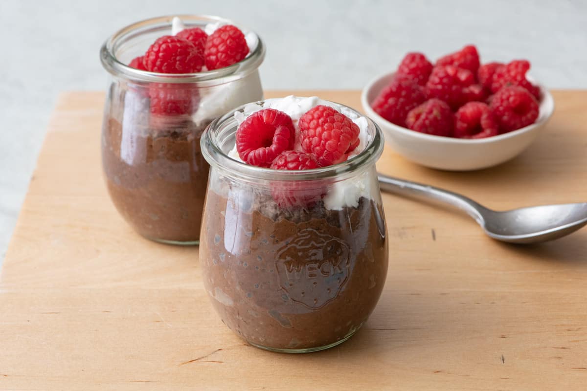 2 small jars of homemade chocolate chia pudding topped with cream and fresh raspberries on a wood surface with a spoon nearby and an extra dish of berries.