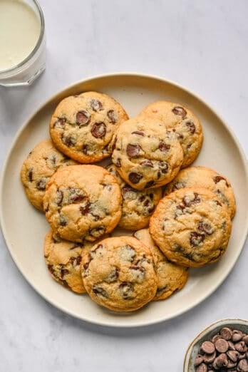 Chocolate chip cookies on a round plate with a small dish of chocolate chips and glass of milk nearby.