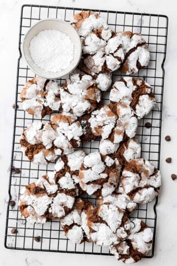 Chocolate crinkle cut cookies on a wire rack with a small bowl of powdered sugar nearby.