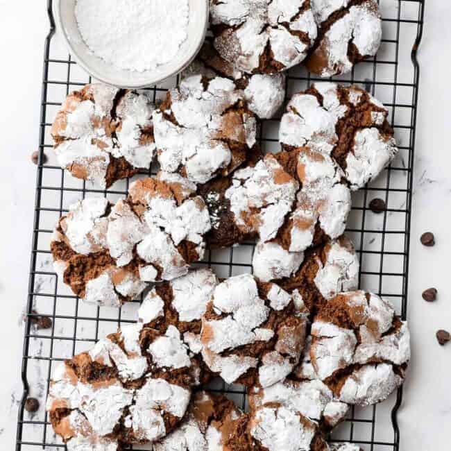 Chocolate crinkle cut cookies on a wire rack with a small bowl of powdered sugar nearby.