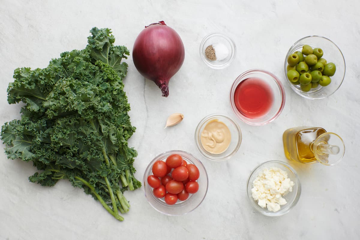 Ingredients for recipe: kale, red onion, cherry tomatoes, garlic, dijon mustard, salt and pepper, red wine vinegar, crumbled feta, green olives, and olive oil.