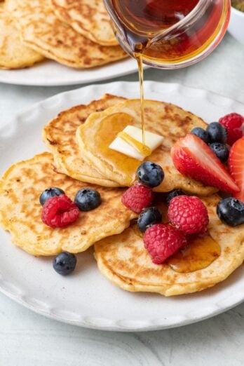 Maple syrup being poured over a plate of cottage cheese pancakes topped with fresh berries and butter with the serving plate of more pancakes nearby.