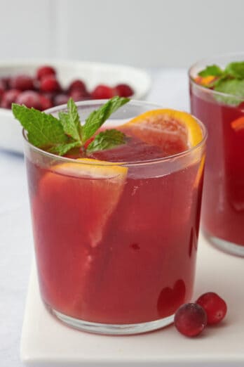 Cranberry orange mocktail in a glass with fresh orange slices and mint with another glass nearby and a bowl of fresh cranberries behind them.