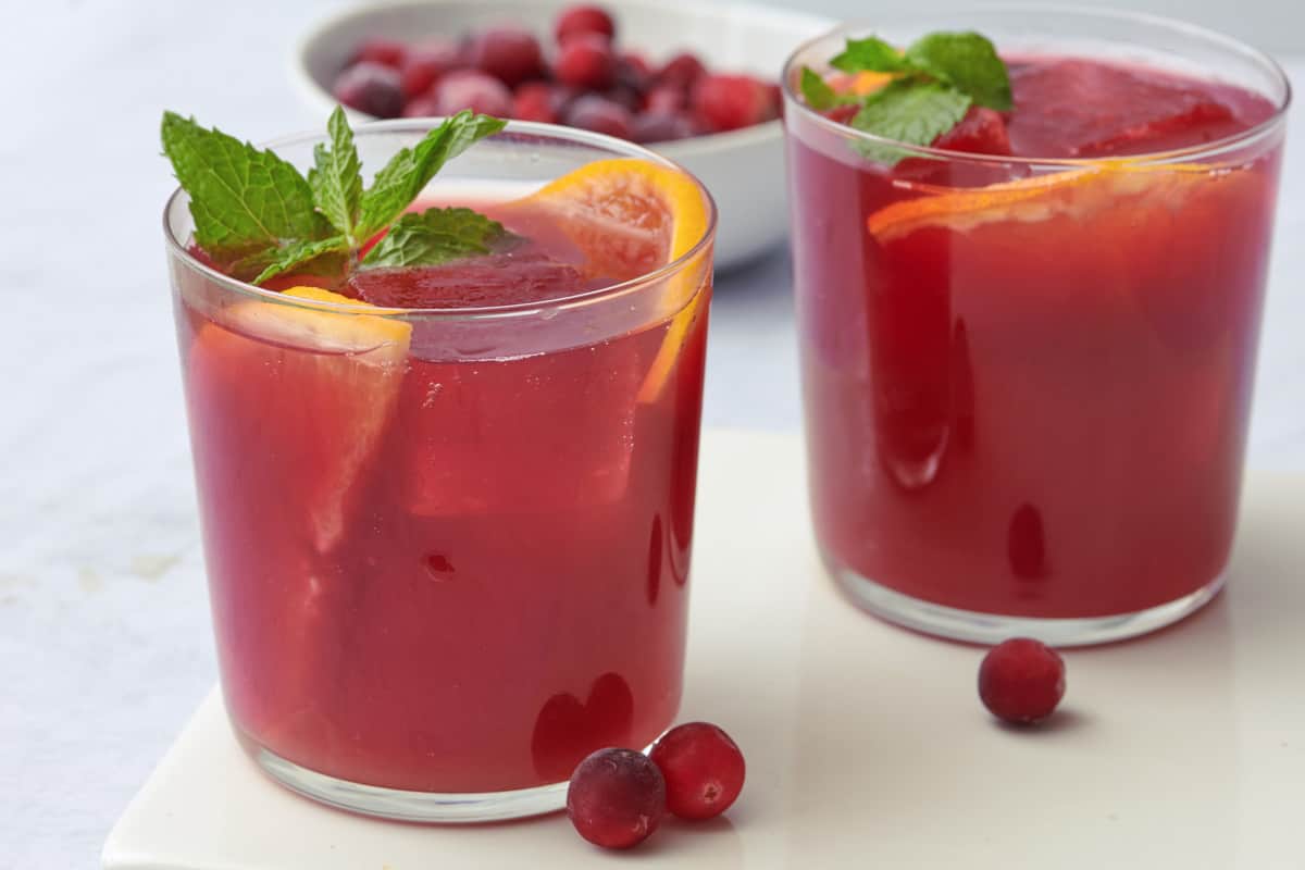 Two glasses of cranberry orange mocktail, both garnishes with orange slices inside and fresh mint leaves. A few fresh cranberries around with a bowl of more in the background.