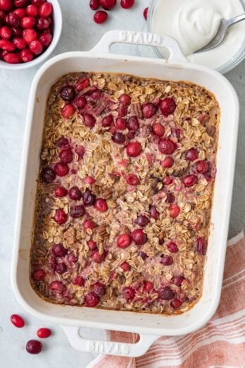Baked cranberry pecan oatmel in baking dish with fresh cranberries and yogurt nearby in small bowls.