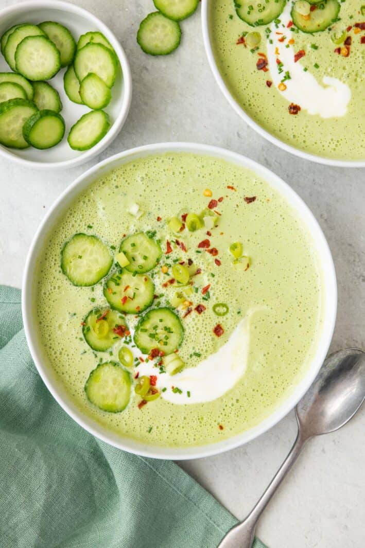 Two bowls of cucumber gazpacho with focus on one, garnishes with extra slices of cucumber, red pepper flakes, and a drizzle of thin yogurt.