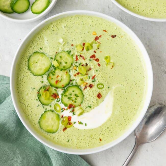 Bowl of cold cucumber gazpacho, garnished with sliced cucumbers, red pepper flakes, and green onions.