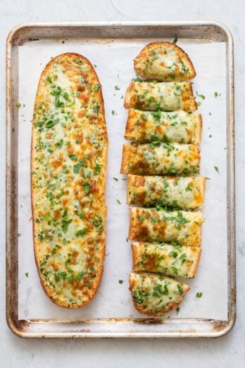 Two baked garlic cheese bread on parchment paper line baking sheet with one bread cut into slices.