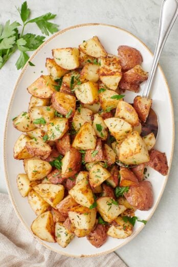 Long serving dish of garlic roasted potatoes garnished with fresh chopped parsley with a serving spoon dipped in.