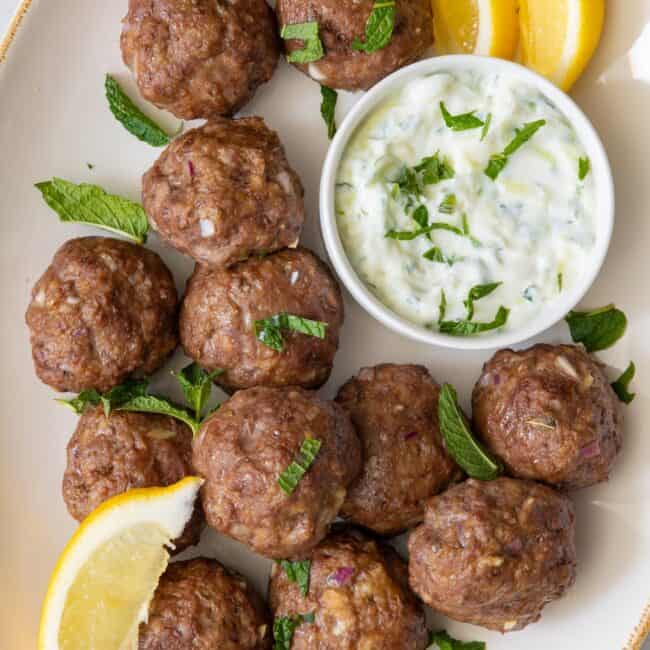 Oval platter with meatballs and side of tzatziki sauce in a small bowl. Garnished with lemon wedges and fresh mint leaves.
