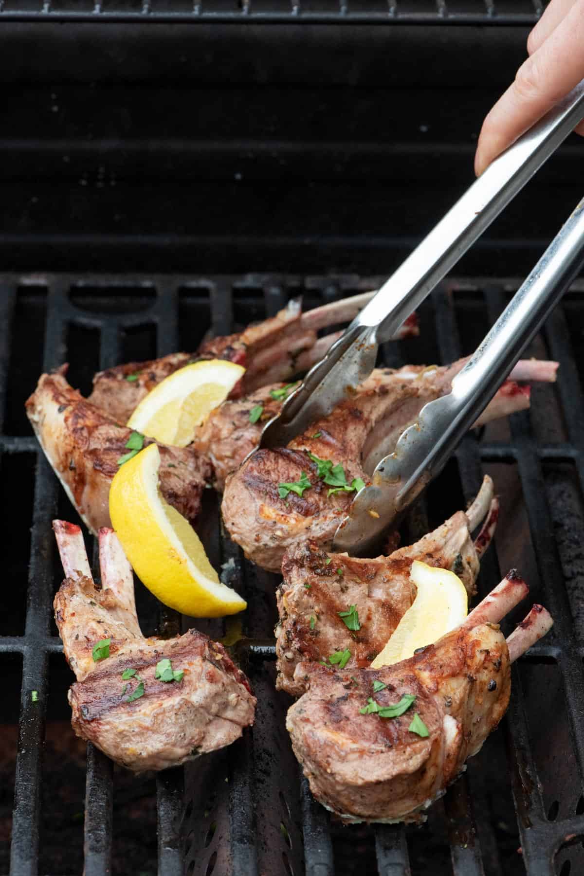 Lamb chops on an outdoor grill garnished with fresh parsley and lemon wedges with tongs lifting one up.