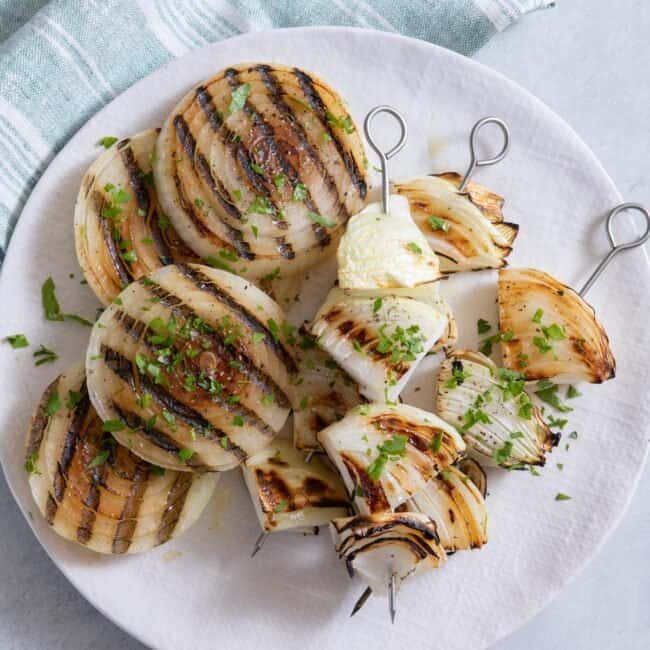 Grilled onion slices and grilled onion chunks on skewers served on a white plate with parley garnish.