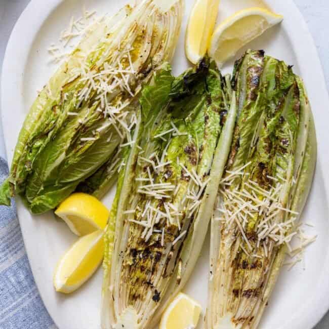 Large white oval platter with 3 halved grilled romaine lettuces sprinkled with shredded parmesan and garnished with lemon wedges.