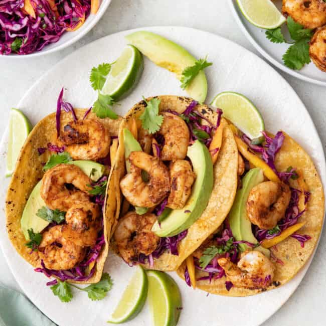 3 grilled shrimp tacos on a plate with sliced avocados, fresh slaw, and lime wedges with a small place of extra grilled shrimp skewers nearby.
