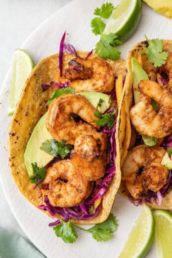 Grilled shrimp taco in a corn shell with a purple cabbage slaw, sliced avocado, cilantro, and lime wedges.