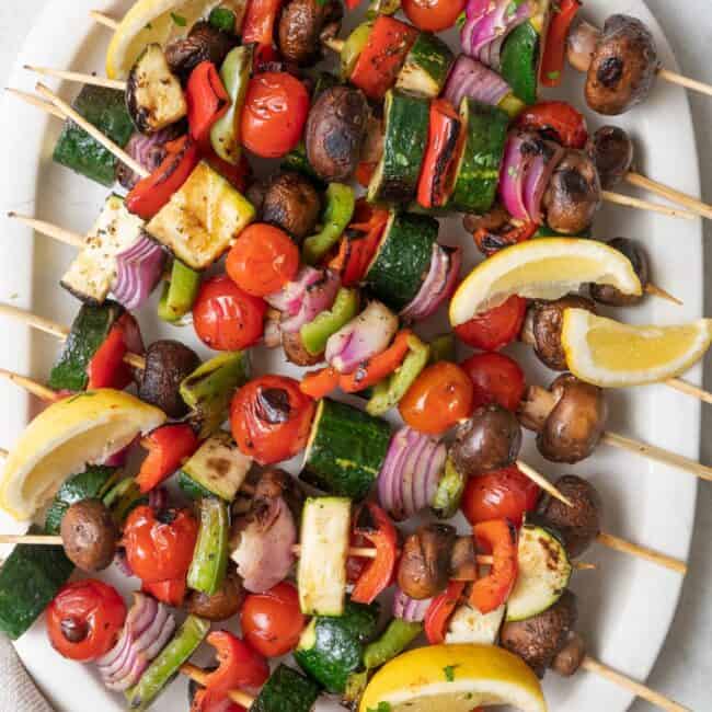 Grilled veggie skewers on a large oval platter garnished with fresh lemon wedges and chopped parsley.