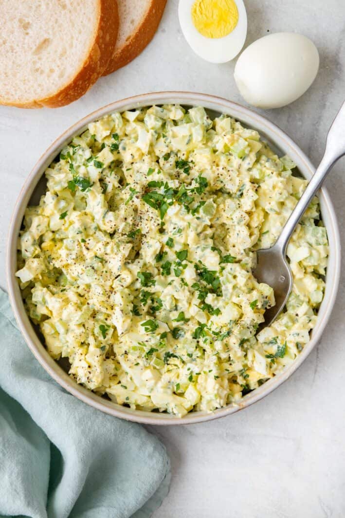 Healthy egg salad in a large shallow bowl, garnished with fresh parsley with sliced bread and boiled eggs nearby.