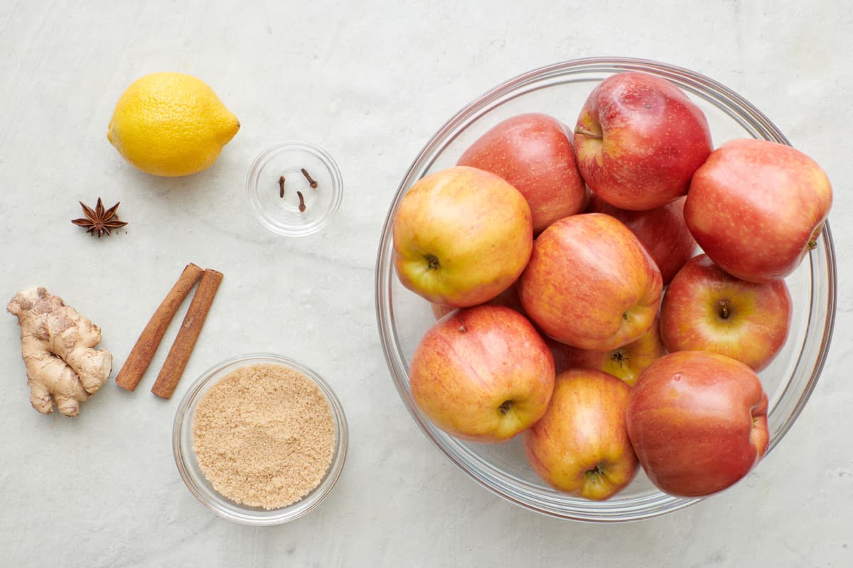 Ingredients for recipe: bowl of apples, lemon, anise seed, clove, cinnamon sticks, thumb of ginger, and light brown sugar.