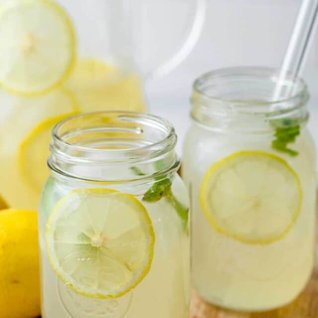 Homemade lemonade in a cup with glass jug in the background and lemons on the side