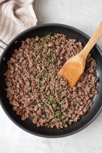 Ground beef in a skillet garnished with fresh parsley and a wooden spatula dipped in.