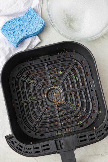 A dirty air fryer basket with cleaning supplies and soapy water nearby.