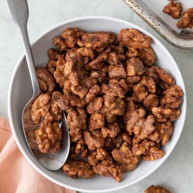 Candied walnuts in a small bowl with a spoon dipped in and baking sheet with more on it nearby.
