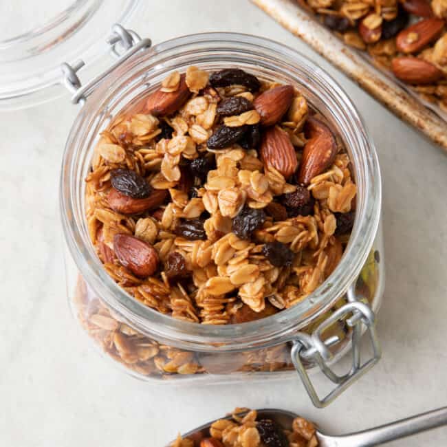 Top down view of homemade granola in an opened small jar with lid, a spoon sitting infront with some on it, and a baking sheet with recipe behind jar.