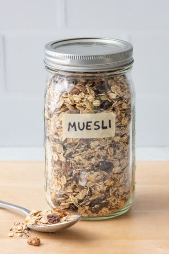 Homemade muesli in a labeled mason jar with the lid on and a spoon nearby overflowing with some.