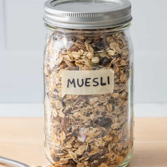 Homemade muesli in a labeled mason jar with the lid on and a spoon nearby overflowing with some.