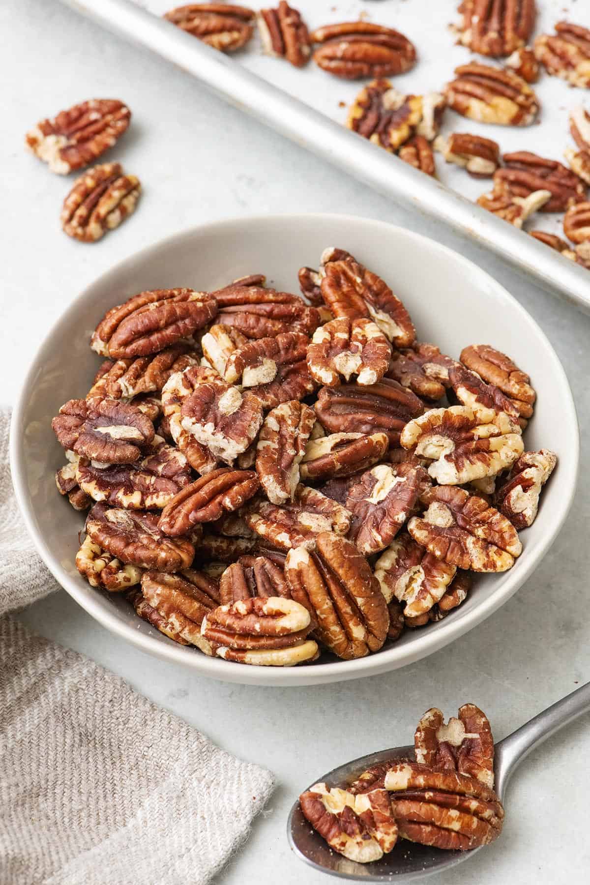 Toasted pecans in a small bowl with baking sheet of more nearby.