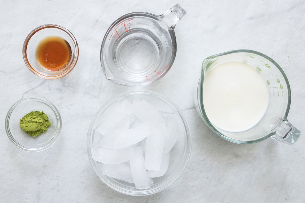 Ingredients for recipe in individual containers: matcha powder, vanilla, hot water, ice cubes, oat milk.